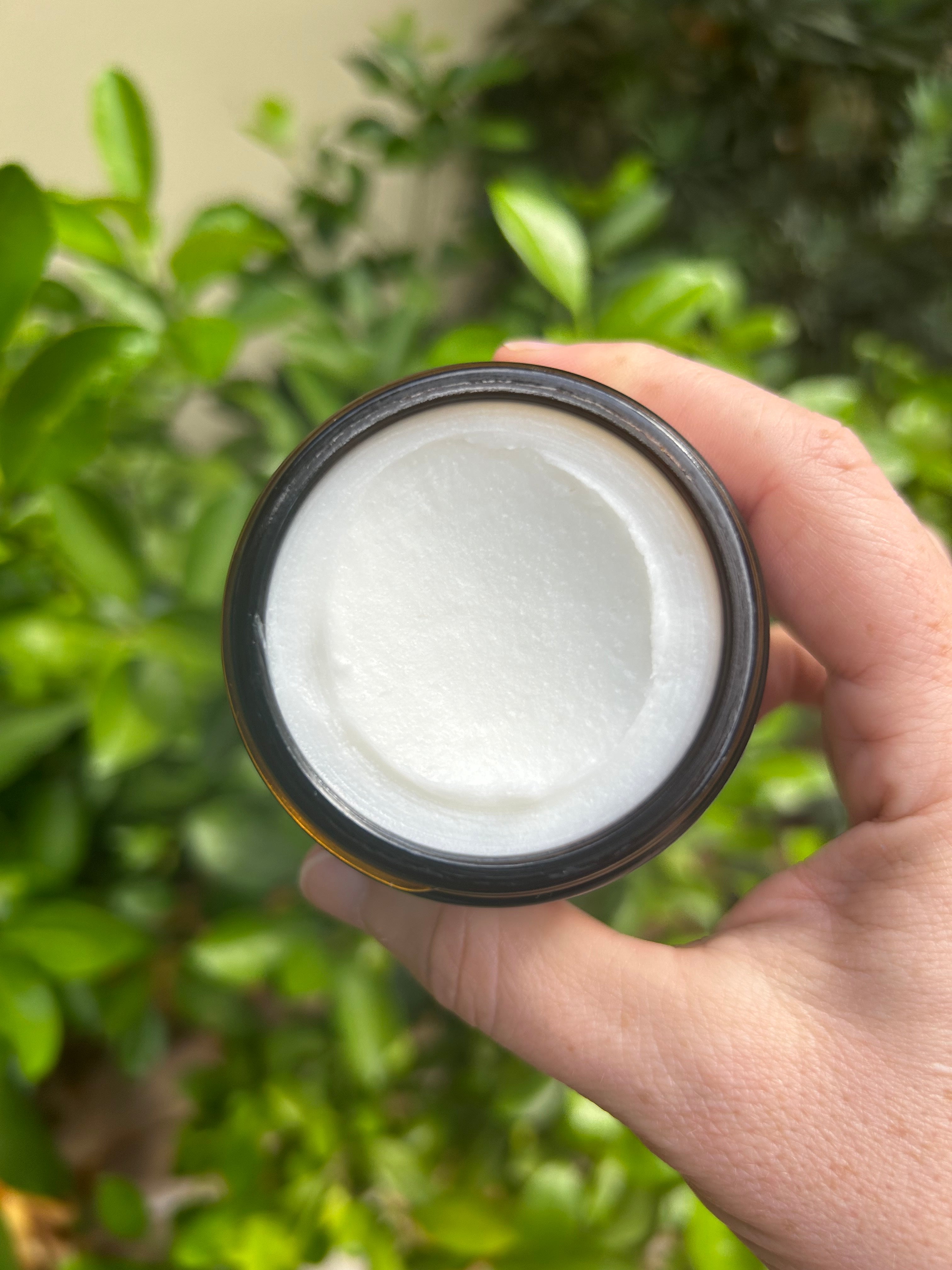 Looking down on an amber glass jar of white body butter with leaves and greenery in the background.