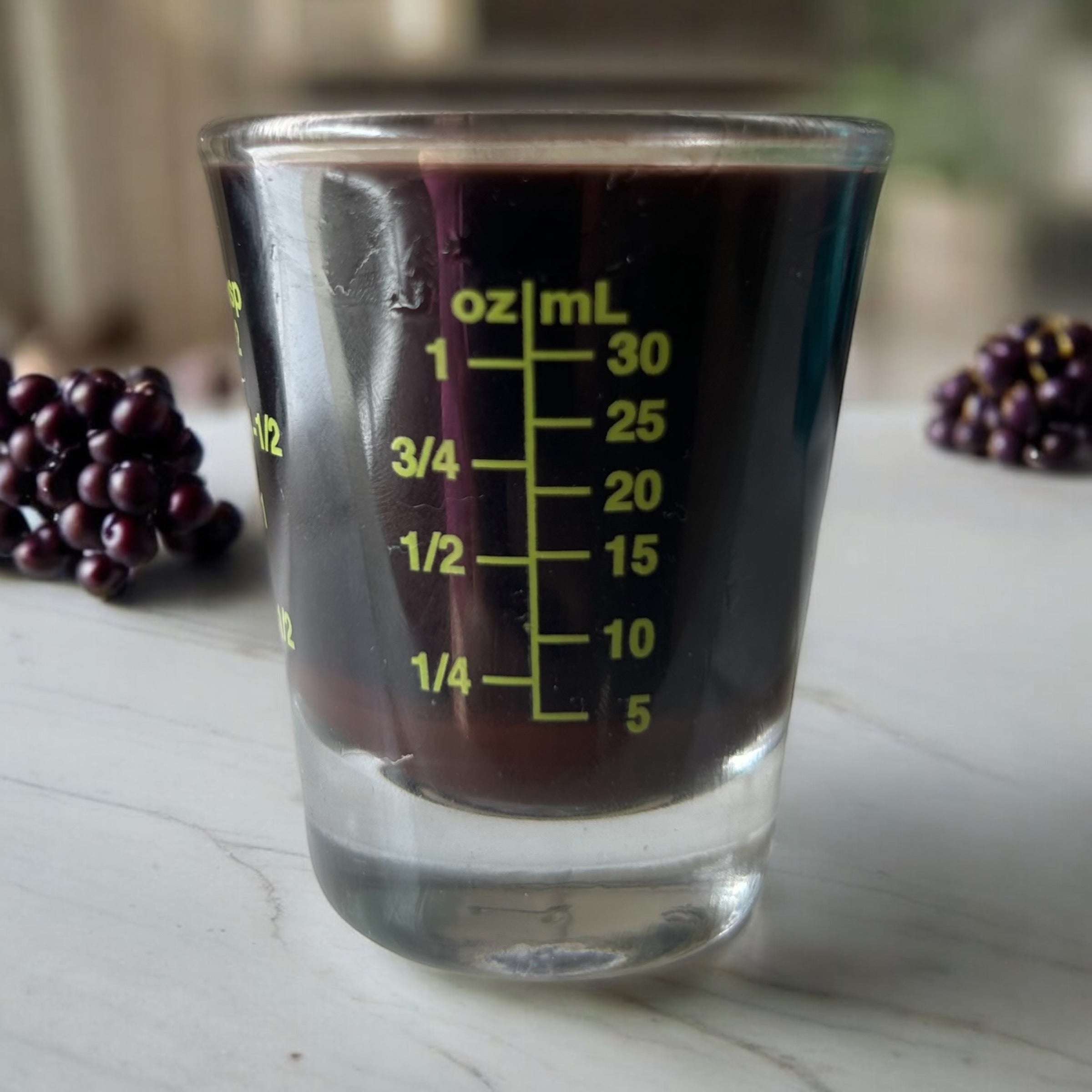 A measuring shot glass with oz/ml in green lettering with elderberry syrup inside and berries in the background.