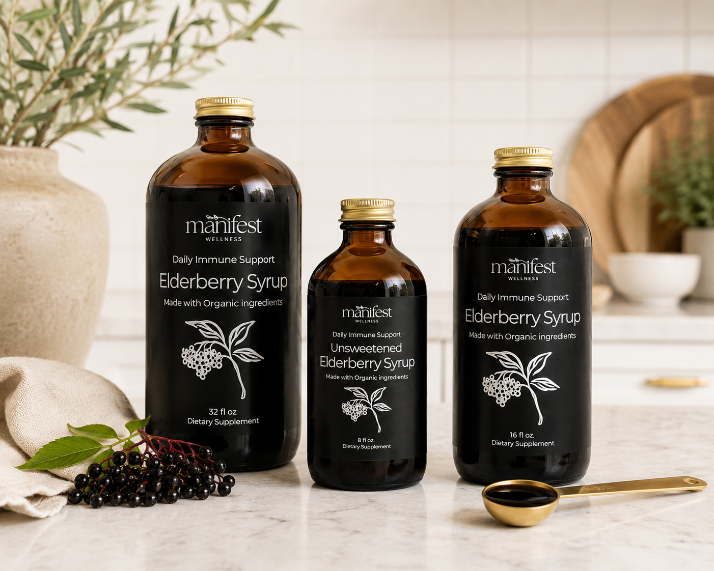 Three amber glass bottles of Manifest Wellness Elderberry Syrup in different sizes, arranged on a light marble surface with a gold spoon and fresh berries in a warm, rustic kitchen setting.