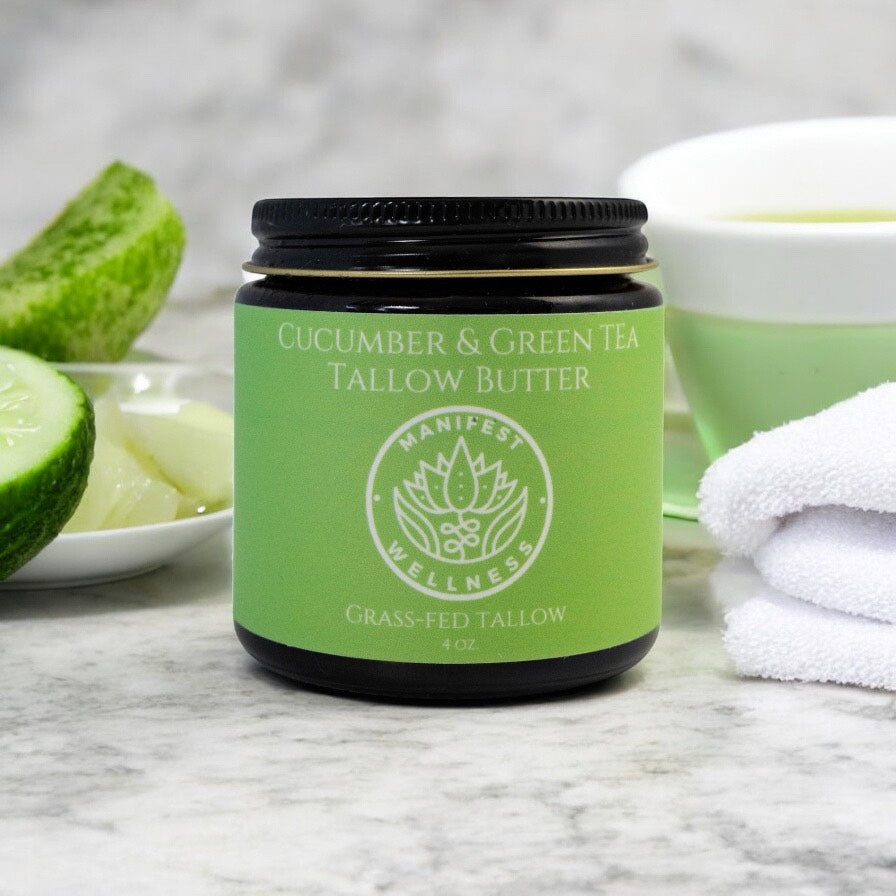 A jar of Cucumber &amp; Green Tea Tallow Butter by Manifest Wellness with grass-fed tallow written on the label. The jar is on a kitchen background with a piece of cucumber and a white towel.