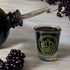 An amber bottle with a stainless steel pourer over a manifest wellness logo shot glass filled with elderberry syrup and berries in the background