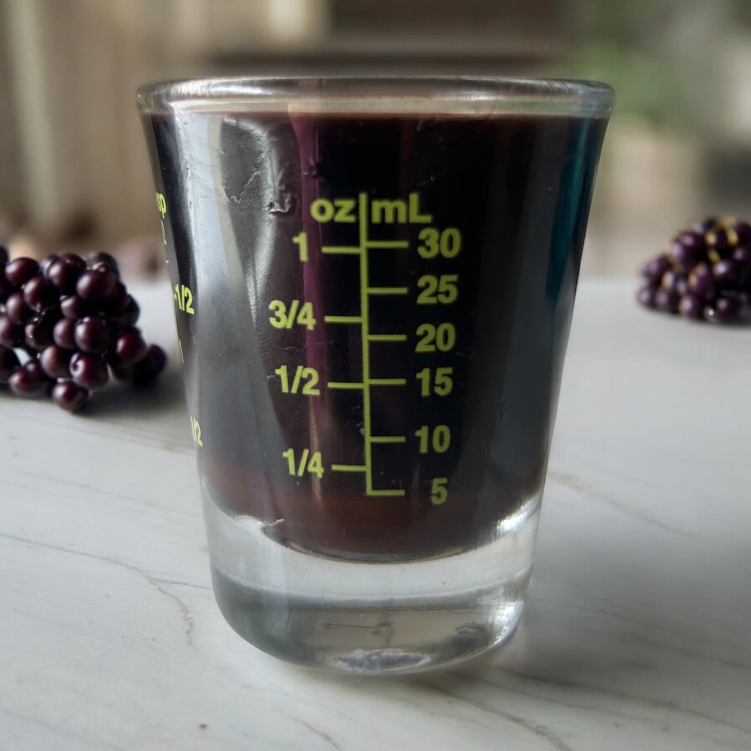 A measuring shot glass with oz/ml in green lettering with elderberry syrup inside and berries in the background.
