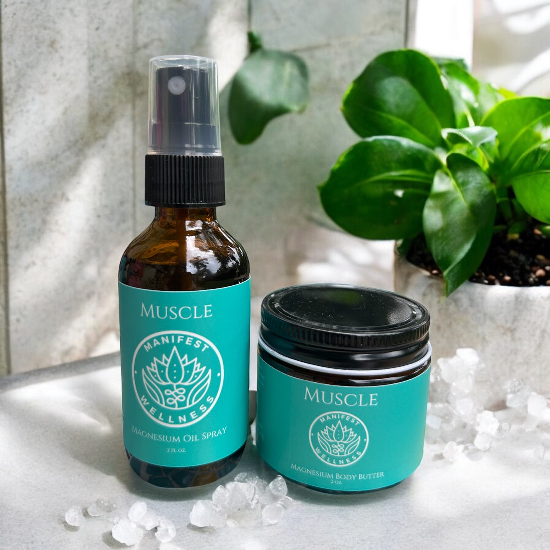 A travel set with 2 oz glass spray bottle and jar of Manifest Wellness Mangesium Muscle Spray and Mangesium Muscle Butter with blue labels. A light bacground with a plant behind and a few magnesium chloride flakes