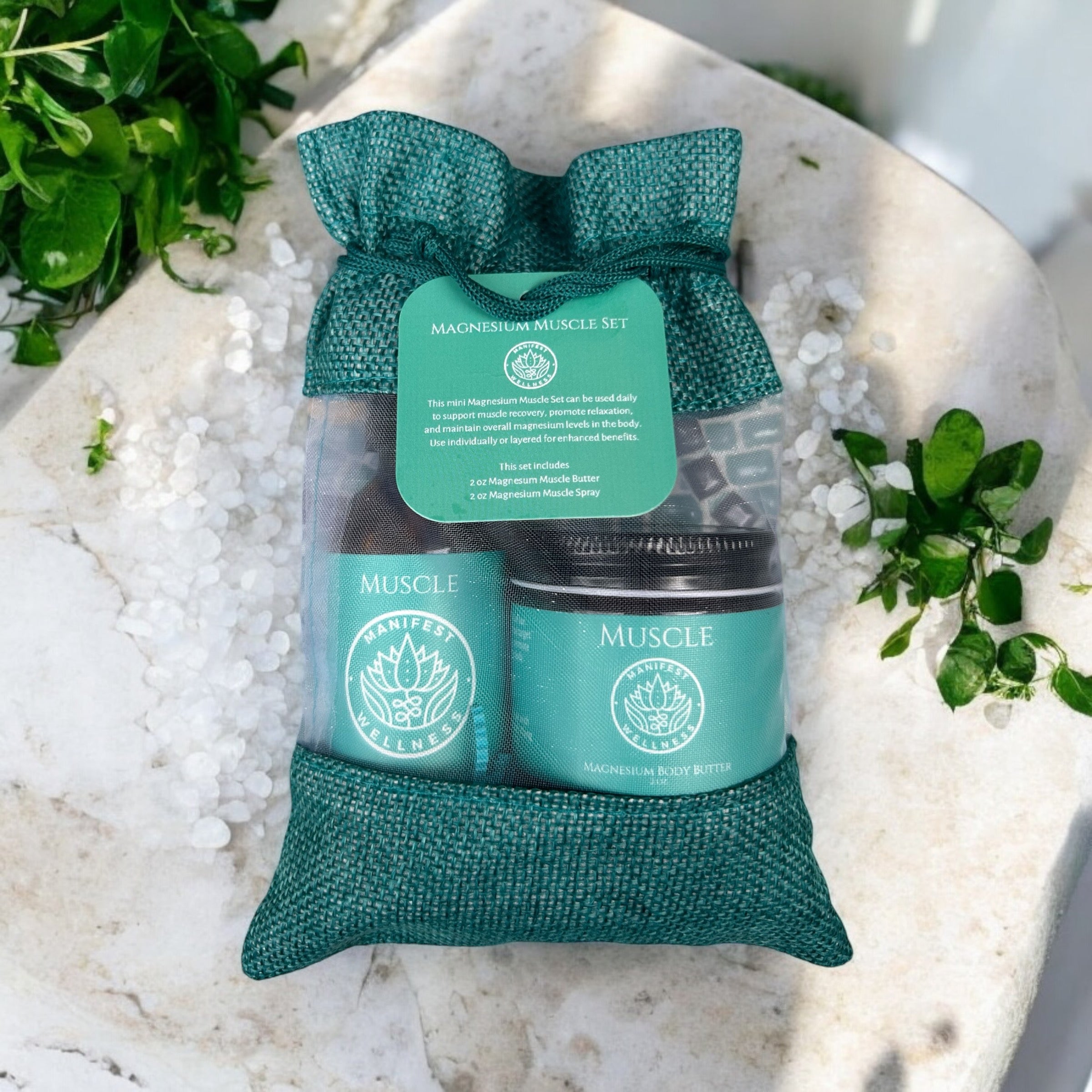 A gift set of Magnesium Muscle Spray and Butter in 2oz glass spray and jar. Products are in a blue fiber bag laying on a table with herbs and magnesium chloride around.
