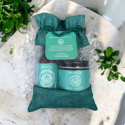 A gift set of Magnesium Muscle Spray and Butter in 2oz glass spray and jar. Products are in a blue fiber bag laying on a table with herbs and magnesium chloride around.