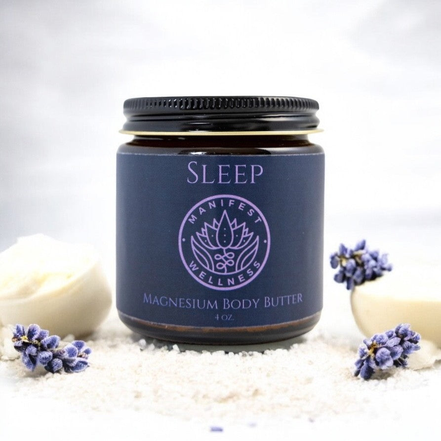 A glass jar of 'Sleep' magnesium body butter from Manifest Wellness with lavender flowers, shea butter and magnesium chloride flakes on a white background