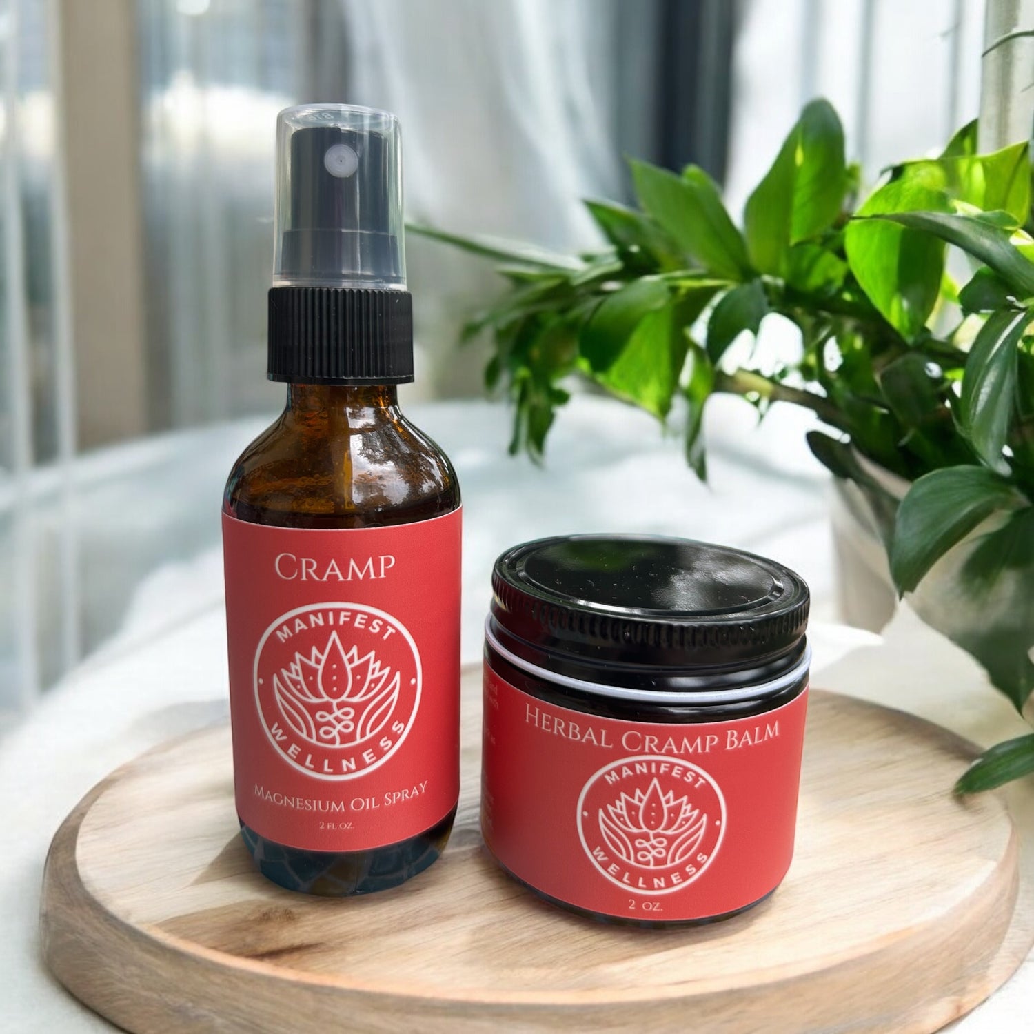 Travel size glass spray bottle and jar with red labels for Cramp Spray and Herbal Cramp Balm in a light background on a wood platform with plants aound.