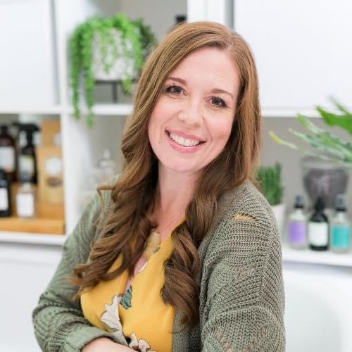 Manifest Wellness owner in a yellow top with a green sweater smiling with arm across a table with glass bottles in the background.