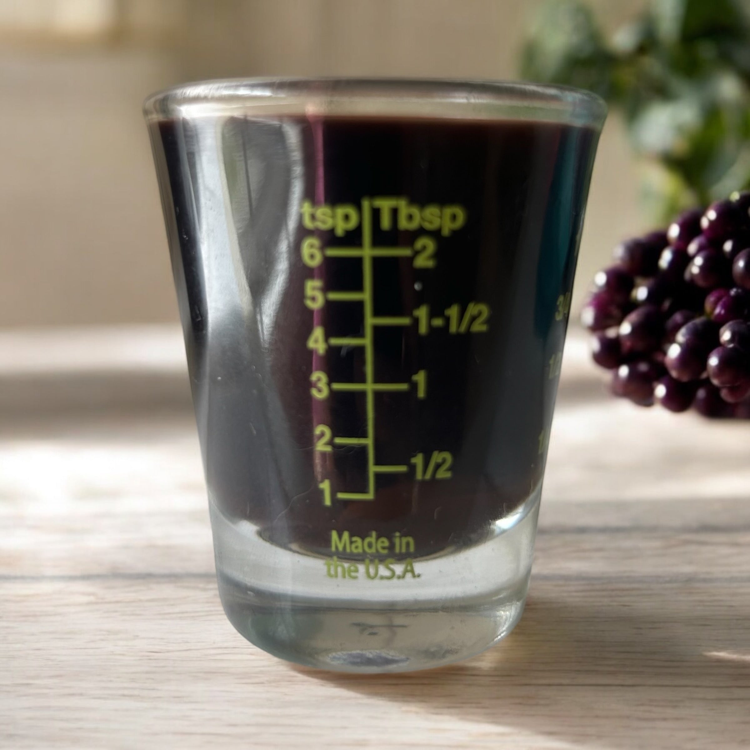 A clear shot glass with green measurements filled with elderberry syrup. On a light wood background with berries behind. 