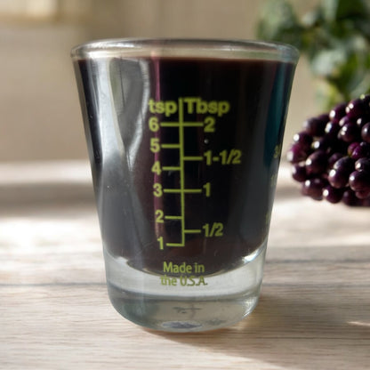 A clear shot glass with green measurements filled with elderberry syrup. On a light wood background with berries behind. 